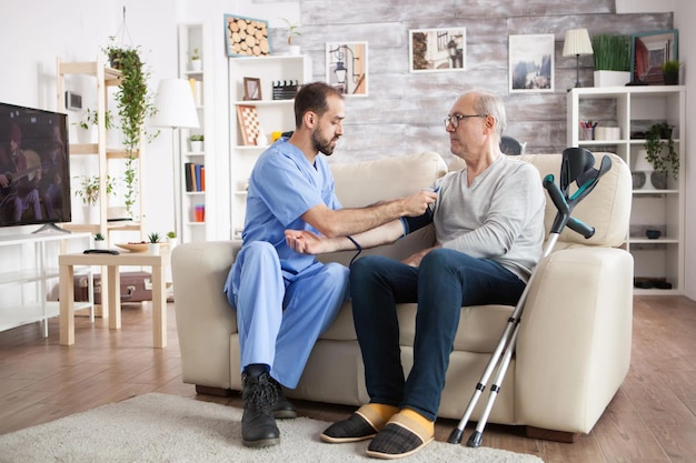 caucasian-young-doctor-nursing-home-checking-blood-pressure-elderly-age-male-with-crutches_482257-45230